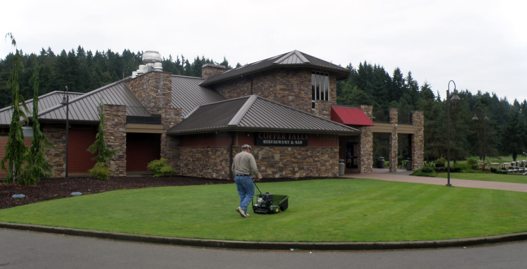 a man walking behind a mower on the yard in front of a restaurant